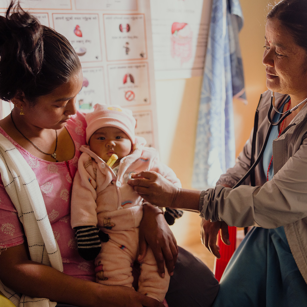 A woman sits on a chair in a medical clinic, holding her baby girl as a female doctor listens to her heartbeat with a stethoscope.